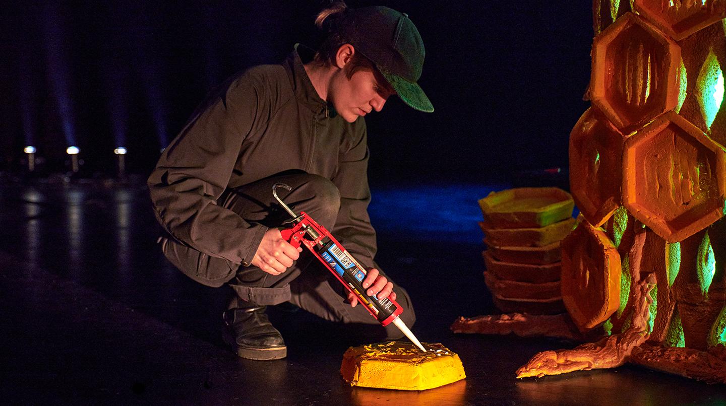 En masterstudent sitter på huk och limmar en scenografidetalj på golvet. Från en scenkonstredovisning 2021. 